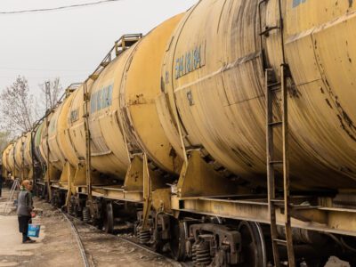 An elderly woman is waiting for a very large oil tanker train to pass so she can cross the street.